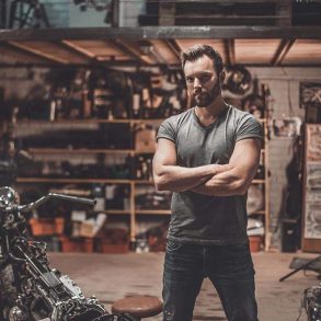 A young man standing next to a motorcycle he is repairing in a garage workshop