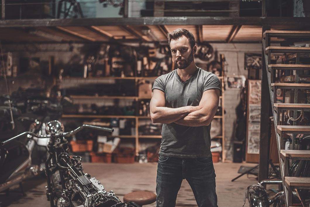 A young man standing next to a motorcycle he is repairing in a garage workshop