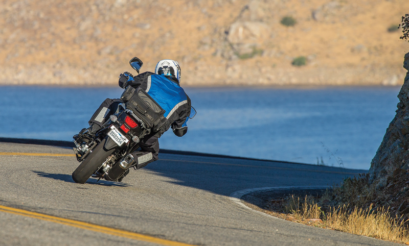 A rider on a motorcycle takes a corner at speed with a lake in the background