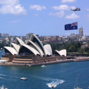 Helicopter with Australian flag flies over Sydney Opera House on sunny day with boats nearby