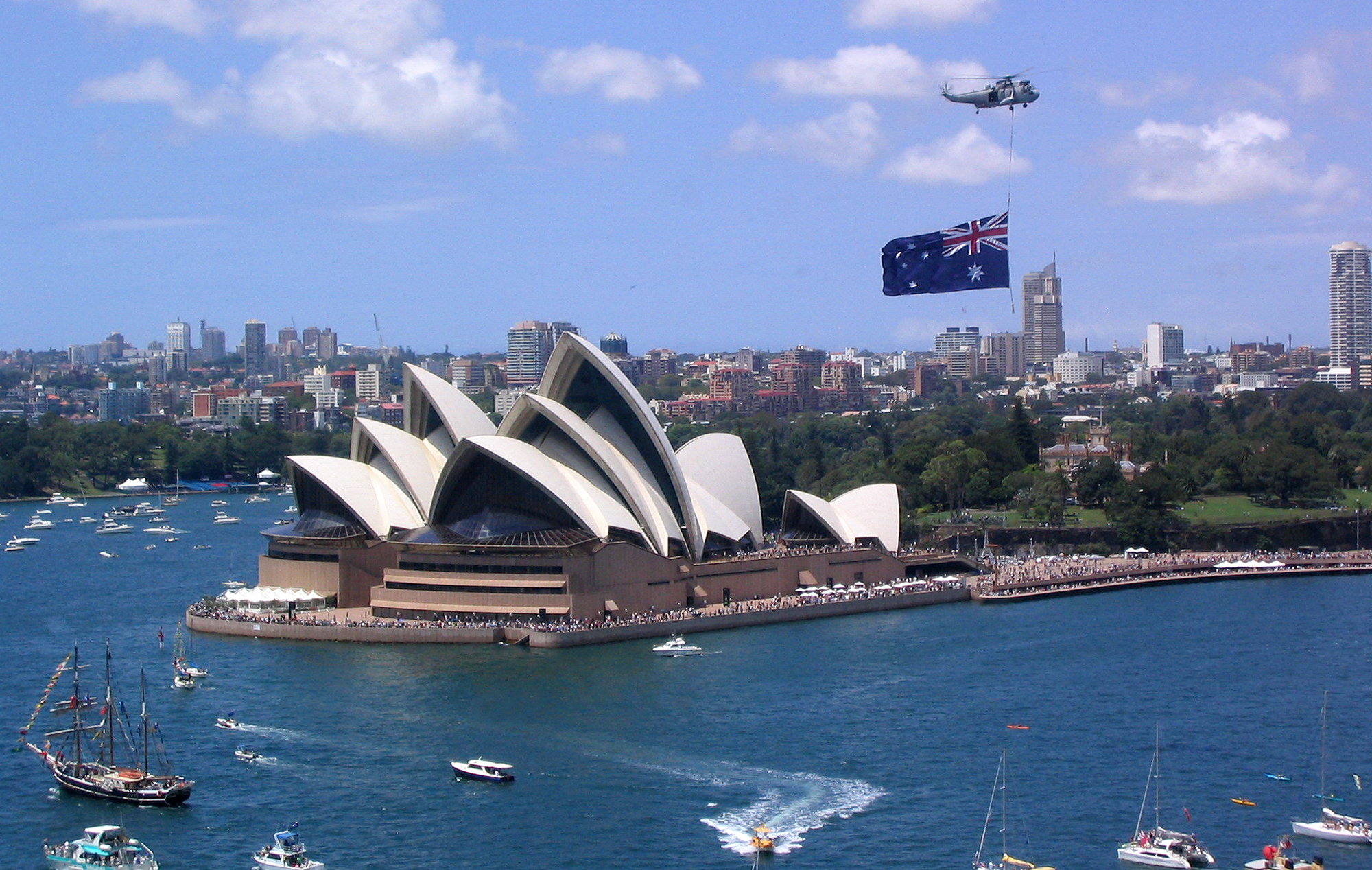 Helicopter with Australian flag flies over Sydney Opera House on sunny day with boats nearby