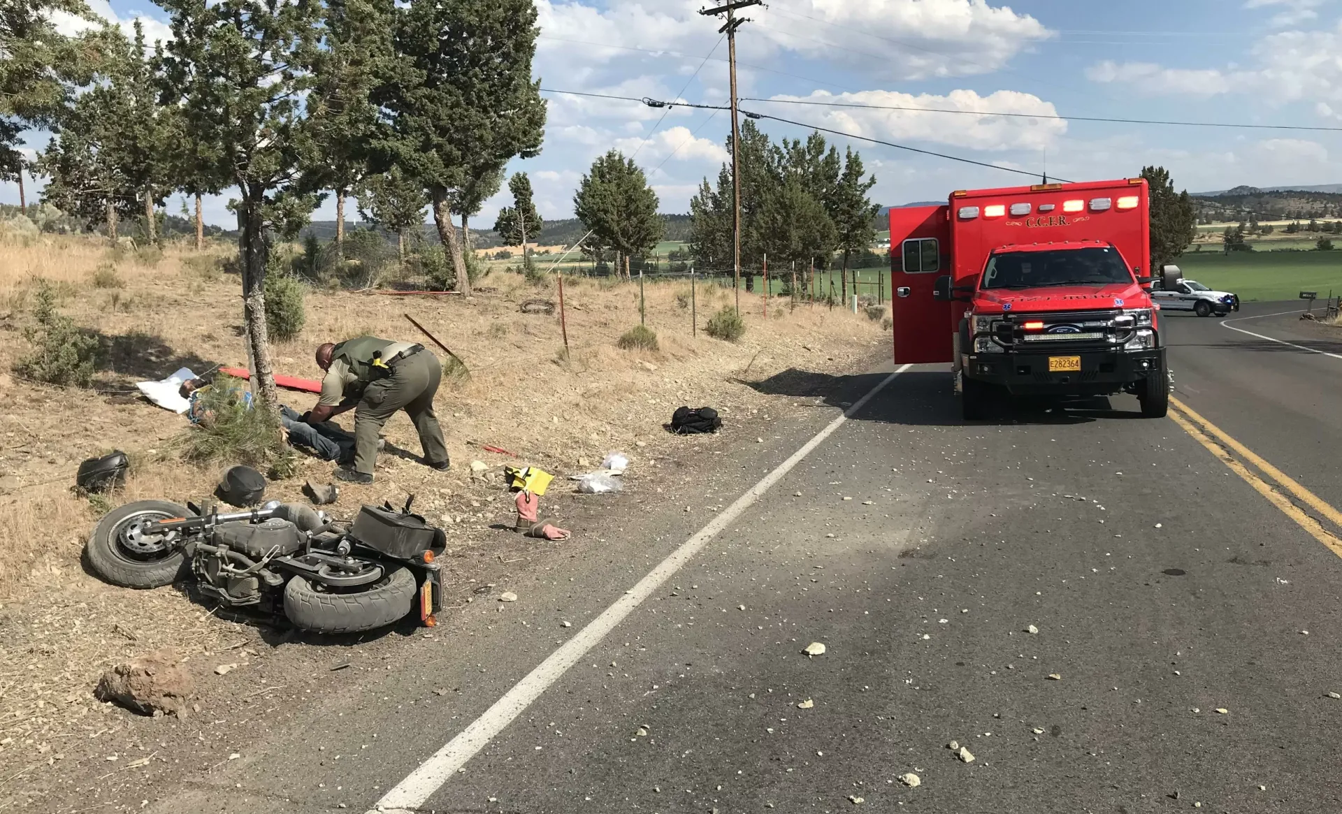 Emergency responders assist an injured person beside a crashed motorcycle on a rural road, with an ambulance nearby.
